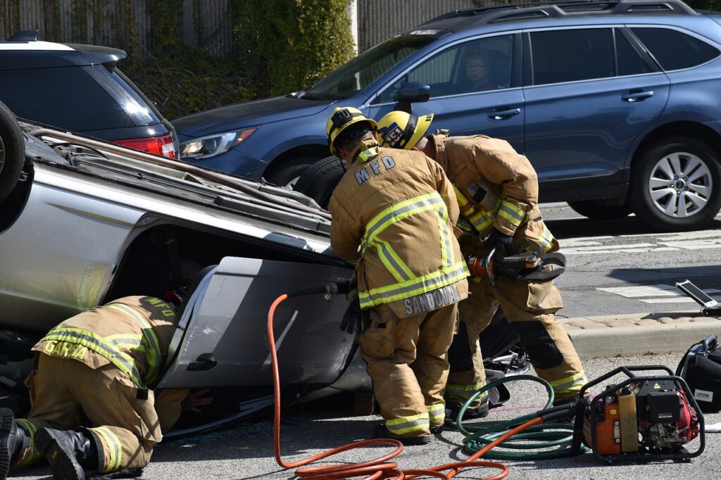 Firefighters rescuing a person from an overturned vehicle after a serious car crash, with emergency equipment and damaged cars visible at the scene.