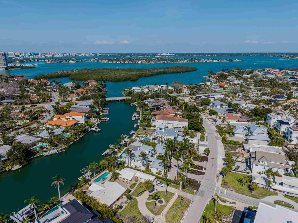 An aerial drone view of a Sarasota, Florida coastal neighborhood showing luxury waterfront homes, palm trees, and residential docks along an emerald green canal leading out to the open bay, indicating the region served by Sarasota personal injury attorneys.