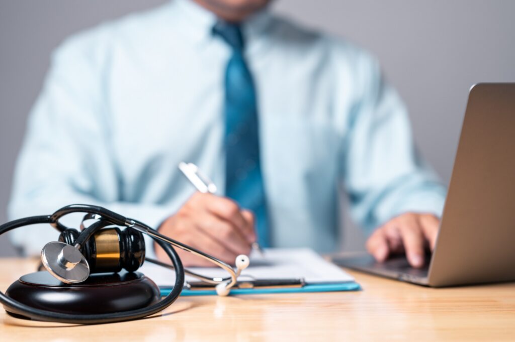 A medical stethoscope and a legal judge's gavel sit on a wooden desk in the foreground, with a blurred professional in a shirt and tie reviewing a clipboard and laptop in the background, representing medical malpractice and injury claims managed by Sarasota personal injury attorneys.
