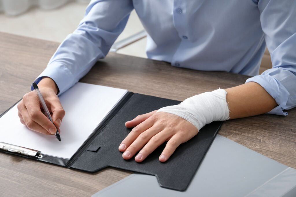 A person in a blue button-down shirt sits at a wooden desk filling out paperwork on a clipboard while their left wrist is wrapped in a white medical gauze bandage, representing a legal consultation with St. Petersburg personal injury attorneys.