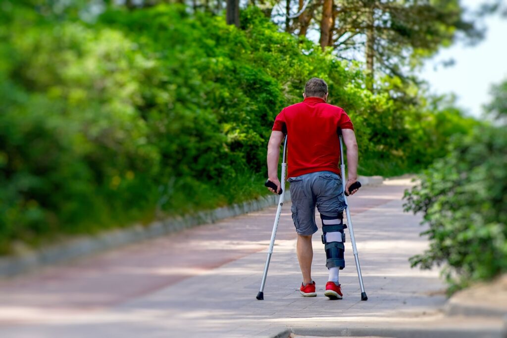 A man seen from behind walking away down a paved outdoor path using forearm crutches and wearing a medical leg brace on his left leg, illustrating physical recovery supported by Stuart & Treasure Coast personal injury attorneys.