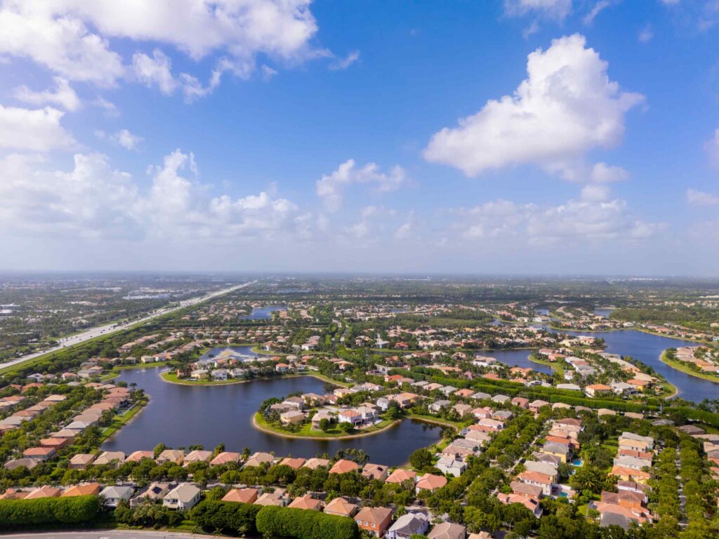 An aerial drone view of a suburban Florida neighborhood in Wellington featuring residential homes surrounding blue lakes and waterways under a bright, partly cloudy sky, indicating the local region served by Wellington personal injury lawyers.